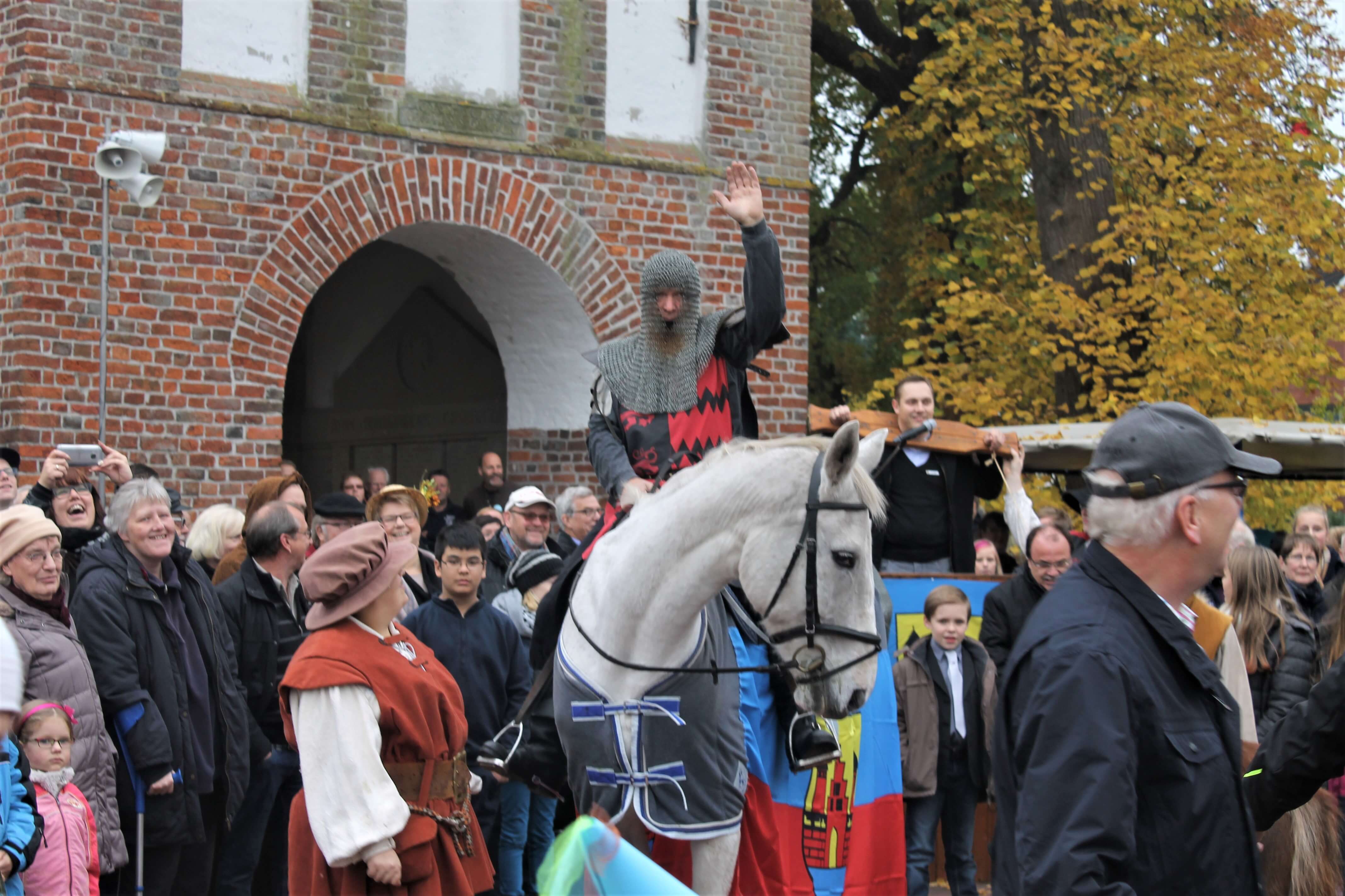 Ein Ritter mit Kettenhaube auf dem Kopf auf einem weißen Schimmel. Im Hintergrund eine begeisterte Menschenmenge.