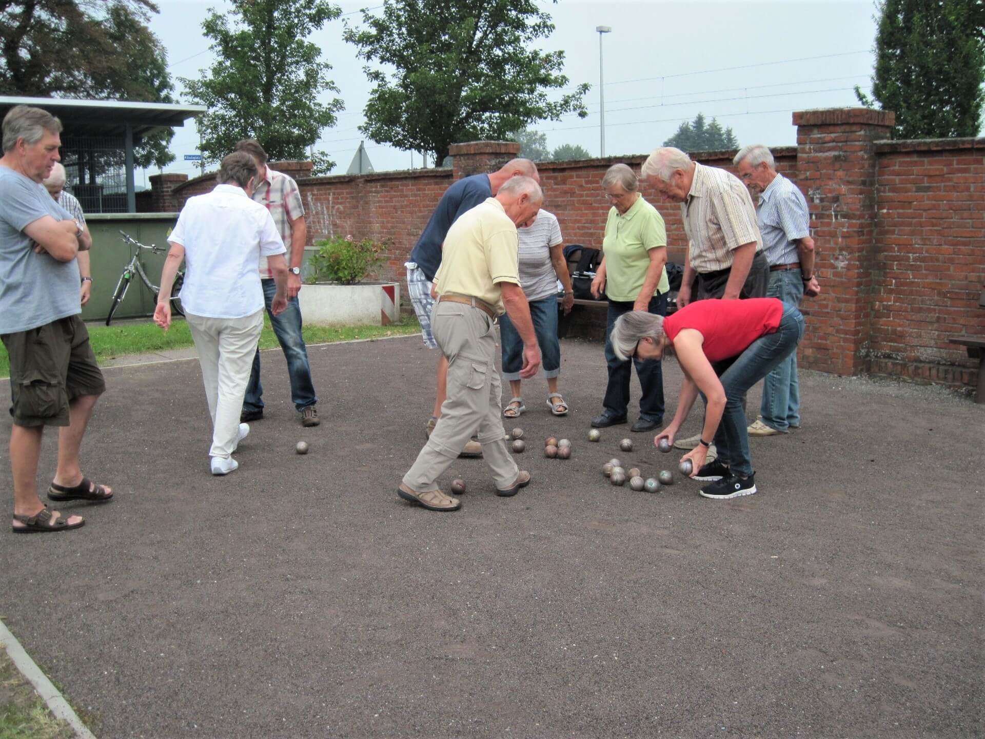 Mehrer Frauen und Männer spielen Boule auf einem Platz neben einer Mauer. Eine Frau in gebeugter Haltung hat zwei Kugeln in der Hand.
