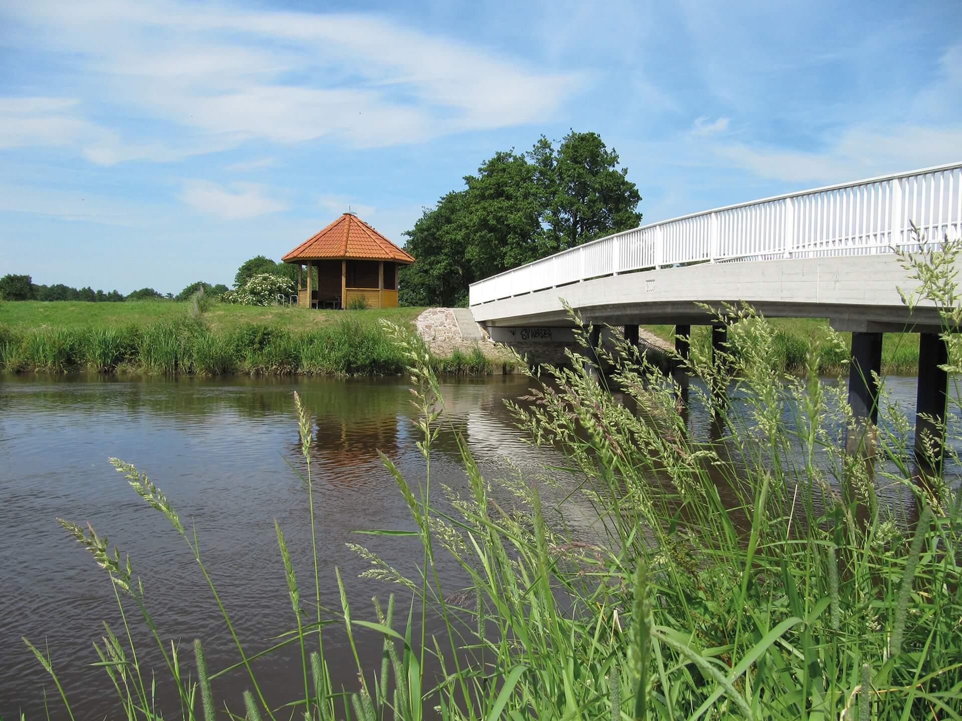 Eine weiße Brücke überspannt einen Fluss. Im Hintergrund steht eine Schutzhütte aus Holz mit rotem Dach