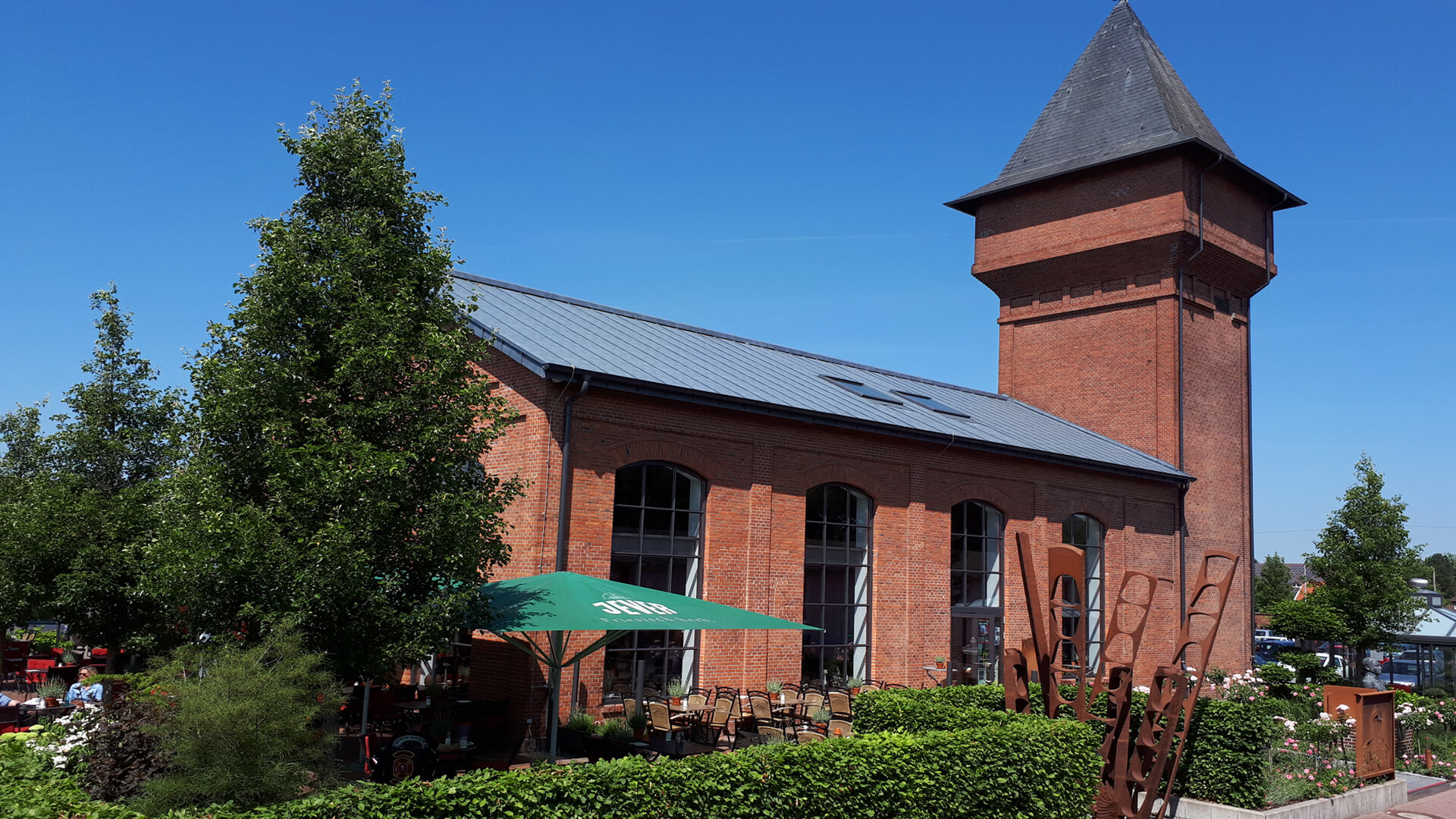 Das Bild zeigt die historische Eisenhütte Augustfehn in Apen, ein rotes Backsteingebäude mit hohem Turm und großen Rundbogenfenstern. Davor steht eine moderne, rostbraune Metallskulptur. Der Garten ist gepflegt, der Himmel blau mit wenigen Wolken – ein sonniger Tag.
