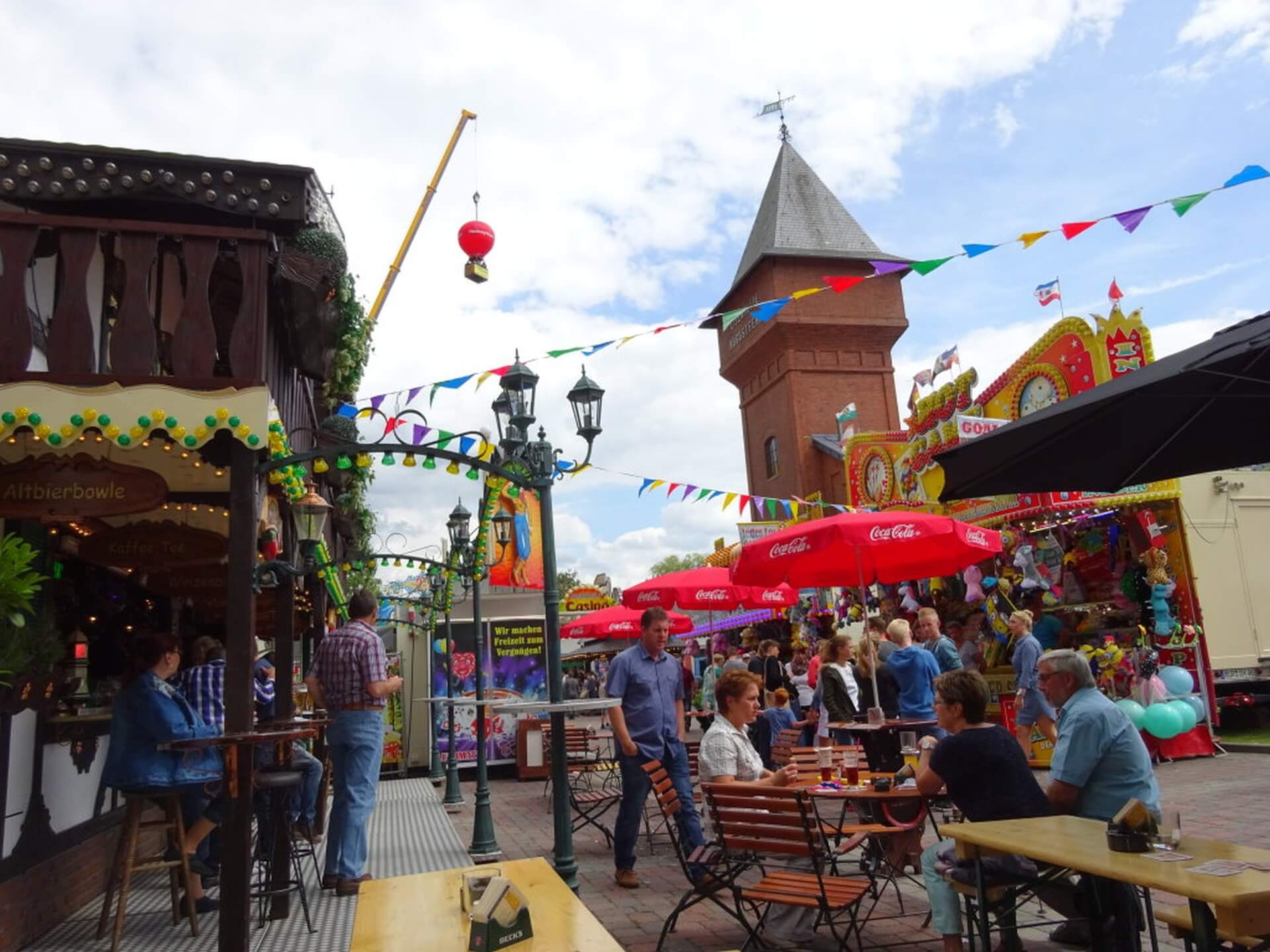 Geschmückter Festplatz mit Menschen, die vor einem Getränkestand auf Stühlen sitzen. Im Hintergrund der Eisenhüttenturm.