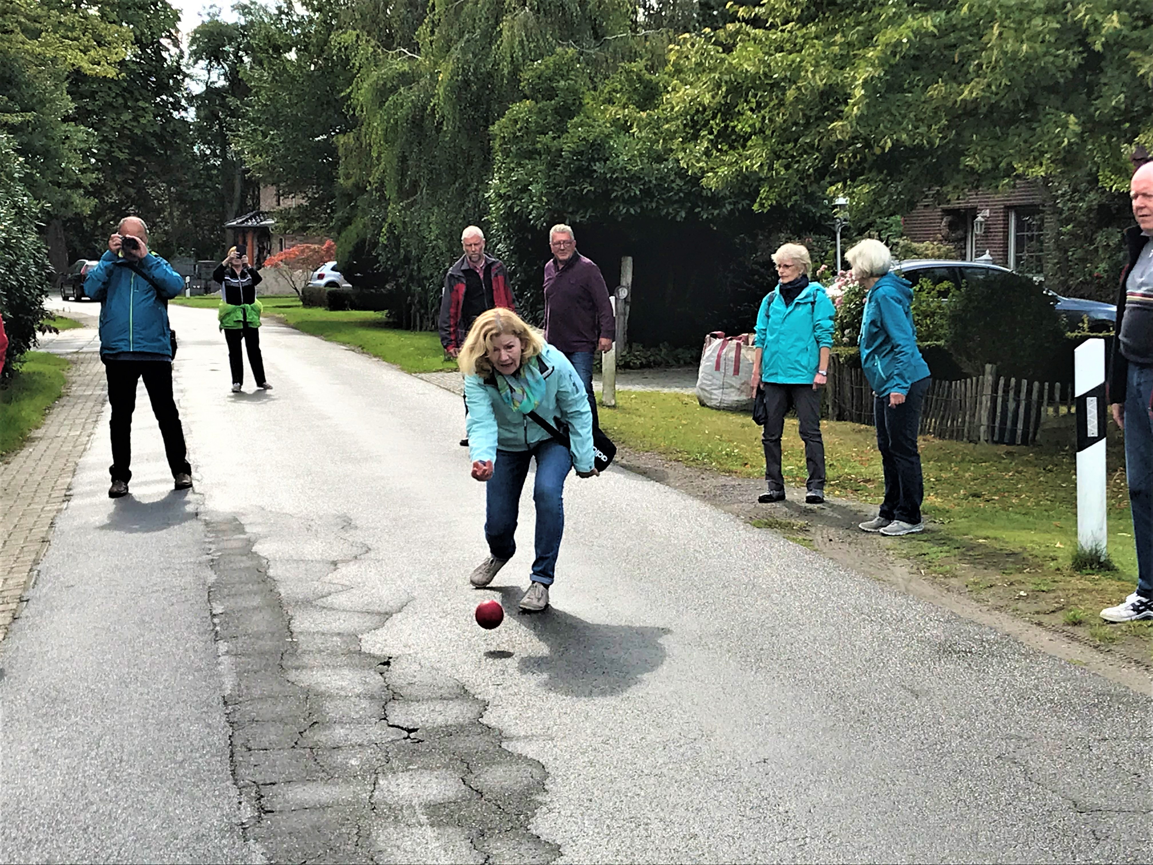 Eine Frau spielt eine Boßelkugel auf einer Straße.Im Hintergrund beobachten und fotografieren die Mitspieler