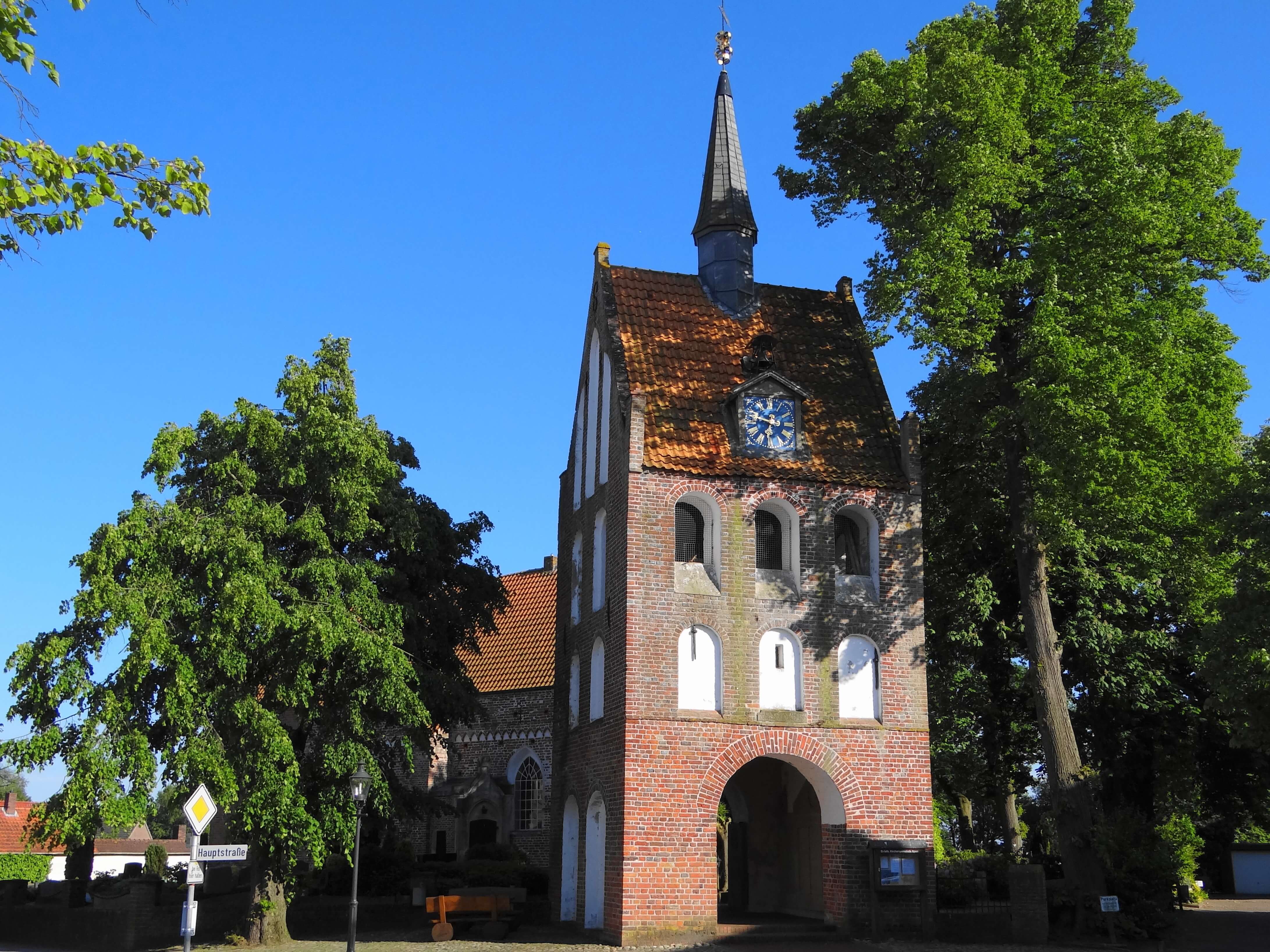 Im Vordergrund steht der prächtige Glockenturm mit vielen weiß verputzten Fensterwölbungen. Im Gibelbereicht befindet sich die Kirchenuhr. Der Himmel ist strahlend Blau. Im Hintergrund belaubter Bäume und die Kirche.