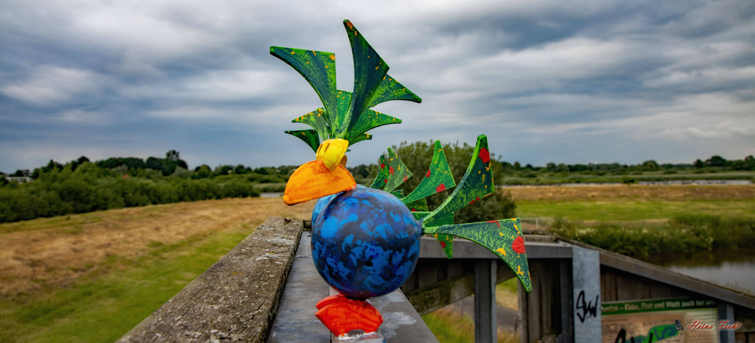 Bunte Vögel aus Stahl sitzen auf einemHolzverschlag. Im Hintergrund ist eine Deichlandschaft