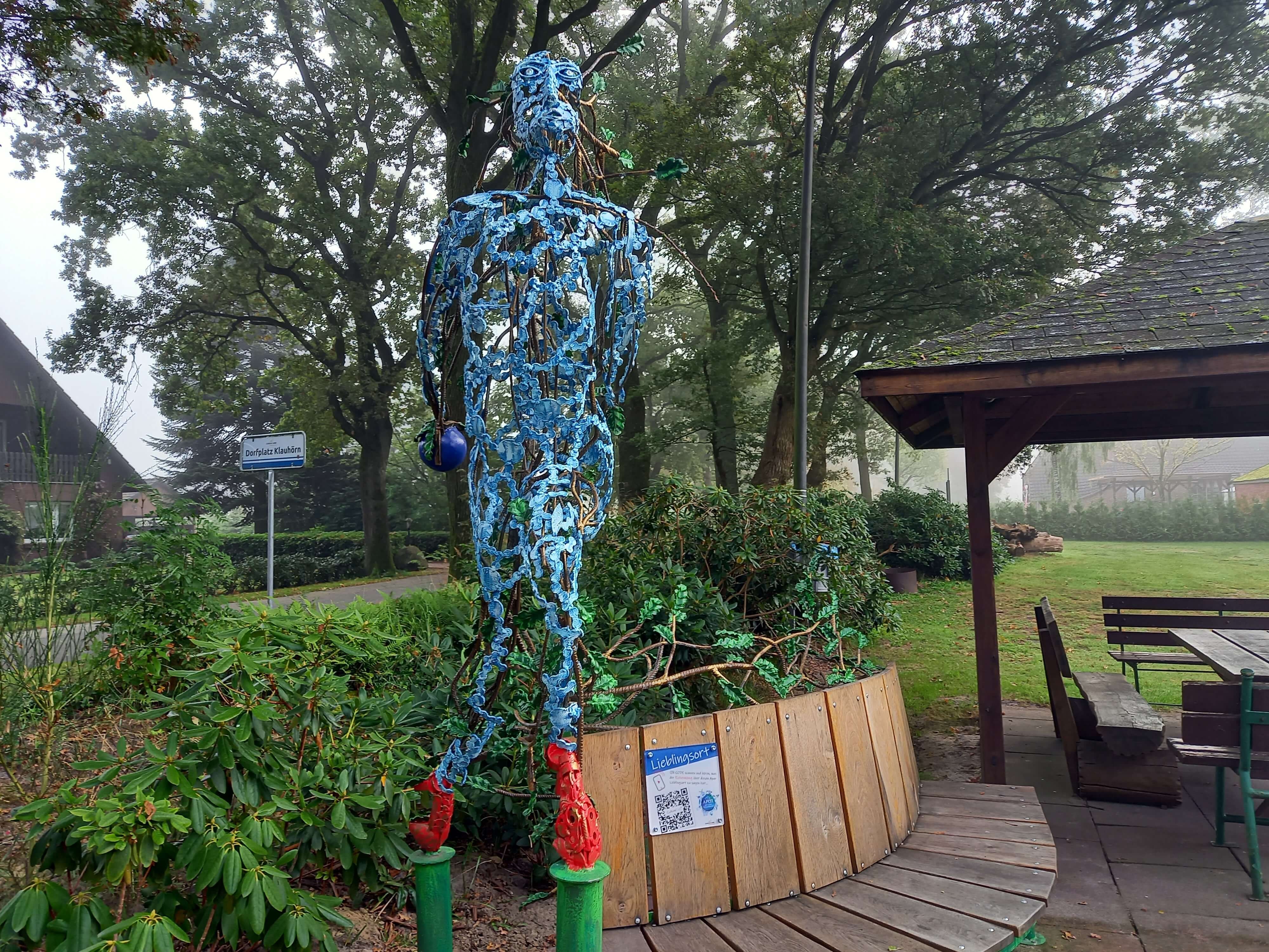 Ein Schild mit einem QR Code ist an einer Holzbank befestigt. Daneben eine bunte Skulptur aus Eisen.