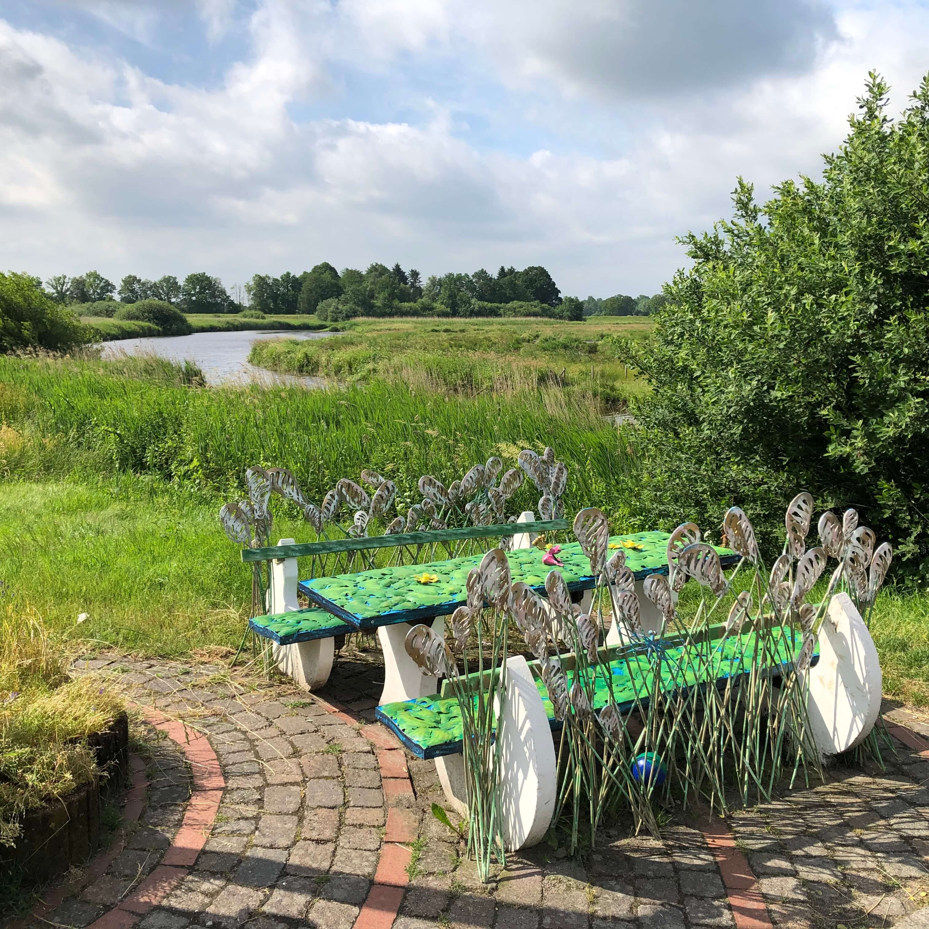 Eine verschiedenfarbige Eisenbank steht auf einem grünen Deich mit Blick auf einen Fluss