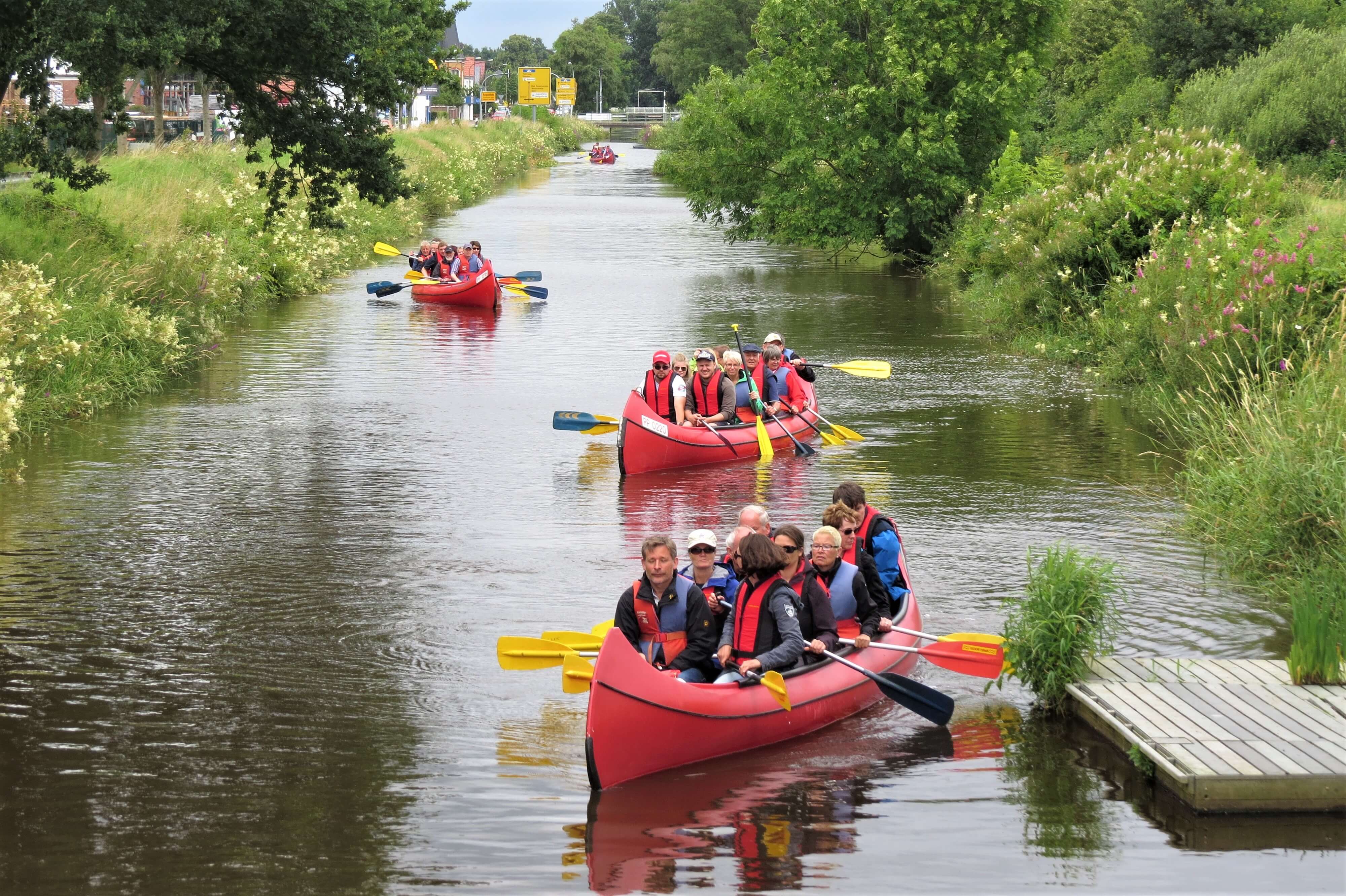 Mehrere Paddelboote auf einem Kanal mit Paddlern in Schwimmwesten legen ab vom Ufersteg. Der Kanal ist eingefasst von grüner Uferböschung.