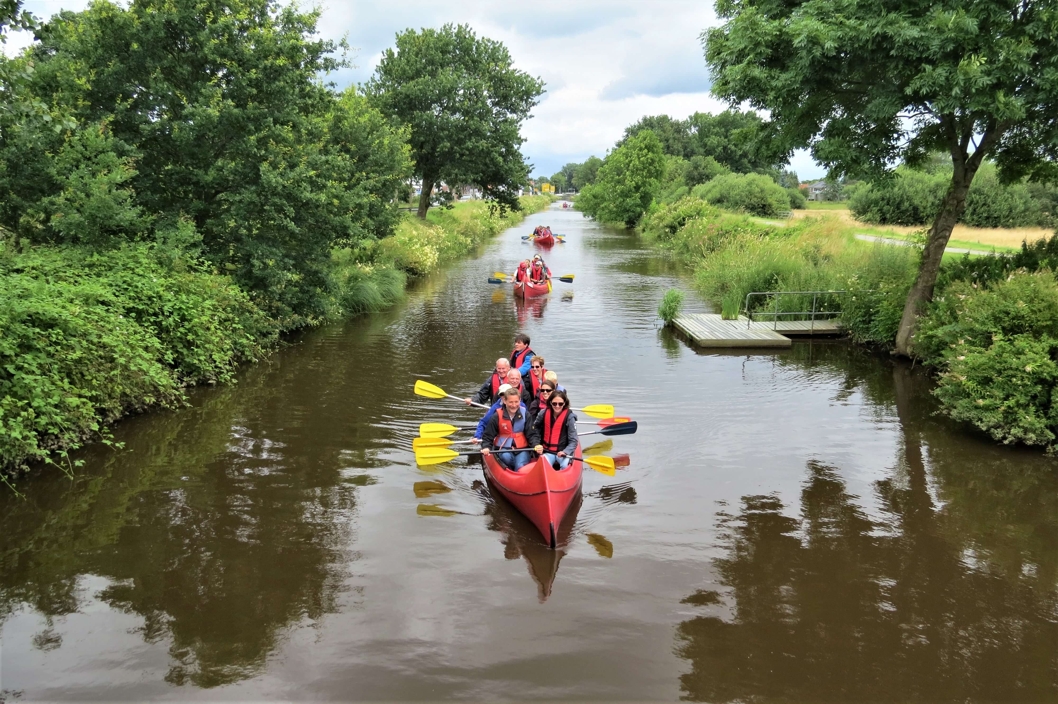 Mehrere Paddelboote mit Paddlern in Schwimmwesten fahren auf einem Kanal mit grüner Böschung am Ufer.