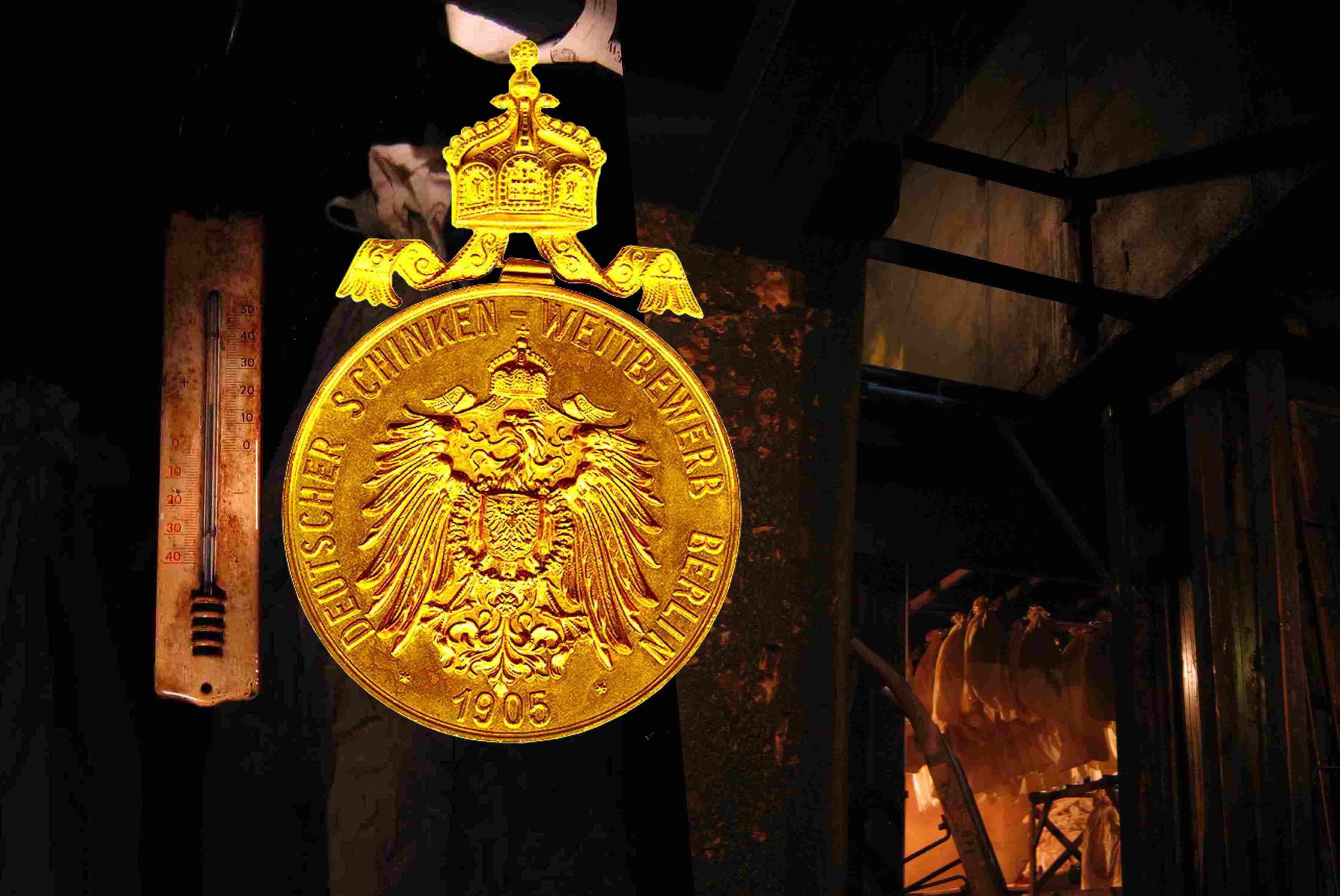 Das Bild zeigt eine goldfarbenen Münze mit dem Titel eines Wettbewerbes aus dem Jahr 1905. Über dem oberen Rand der Münze ist die Abbildung einer Krone mit auslaufend geschmückten Bändern zu sehen. Im Hintergrund befindet sich ein dunkler Raum.