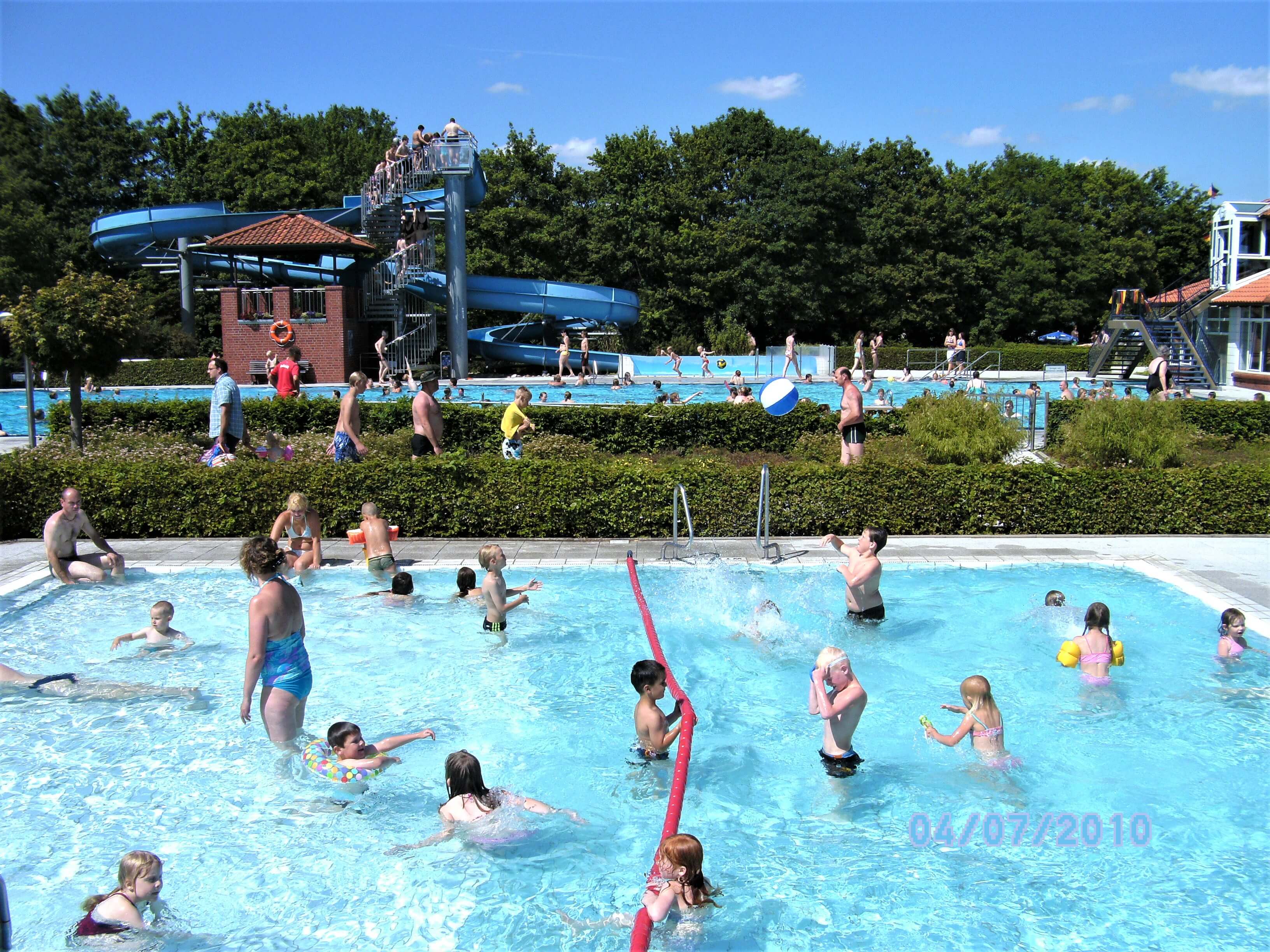 In einem Schwimmbecken mit niedriger Wassertiefe sind mehrere Personen in Badekleidung zu sehen. Im Hintergrund ein großes Schwimmbecken mit einem Turm und einer Wasserrutsche.