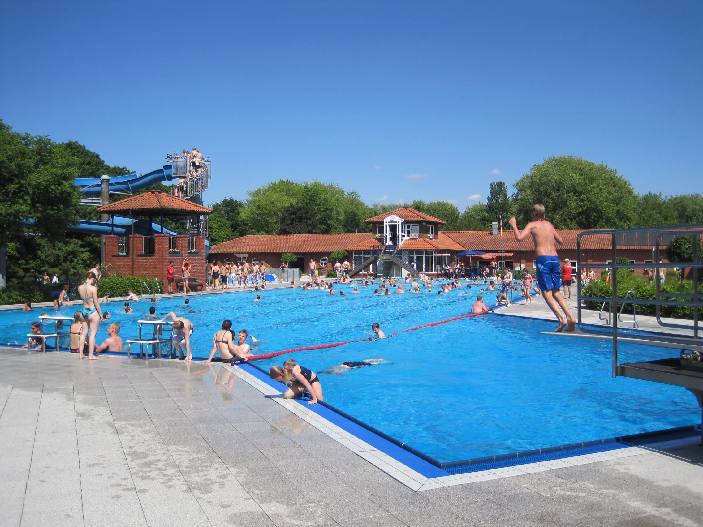 Das Foto zeigt ein Freibad mit viele Menschen in Badekleidung. Im Vordergrund ein Junge, der von einem Sprungbrett in Wasser springt.
