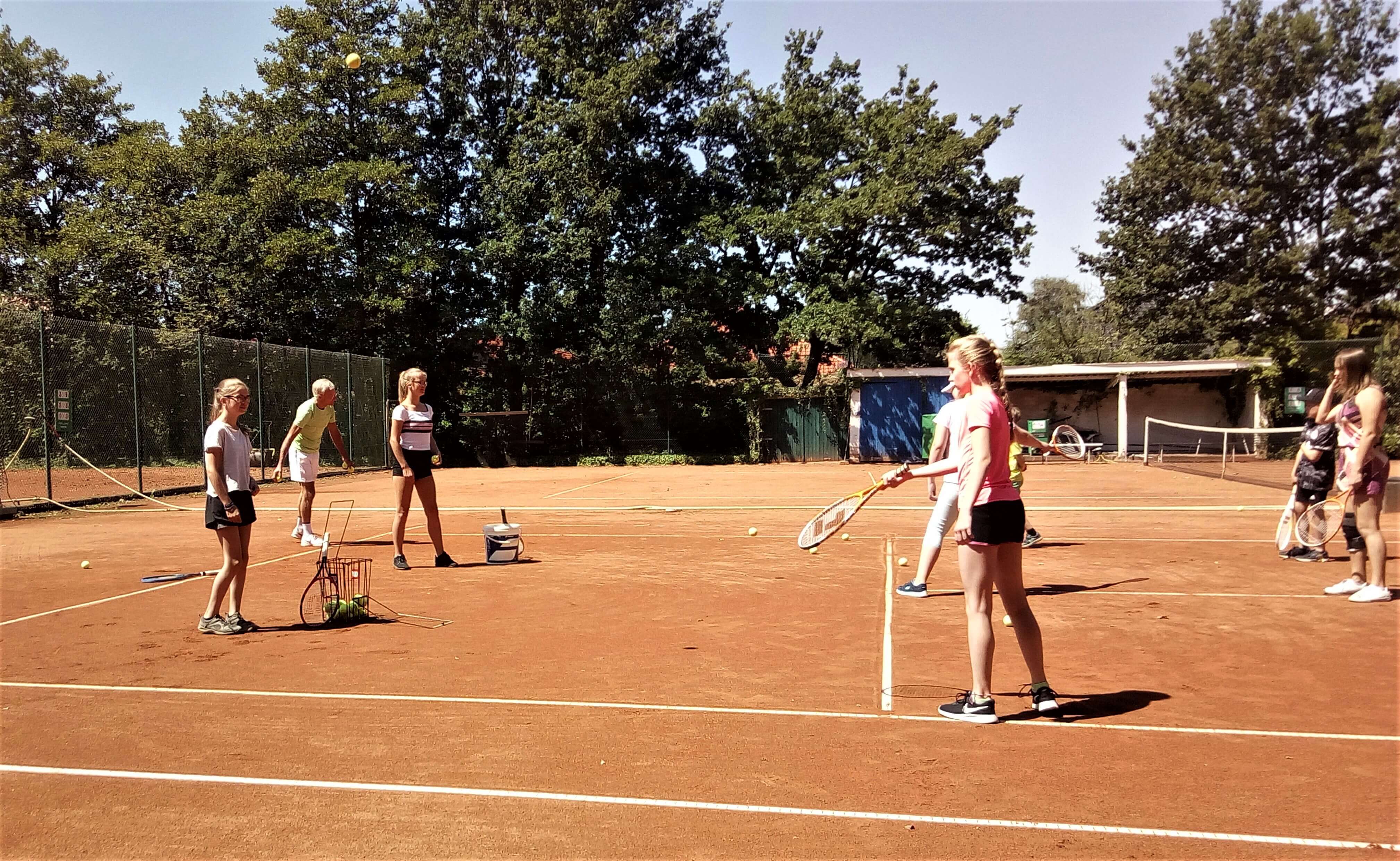 Ein Tennisplatz mit rötlichen Sand im Sonnenschein. Mehrere Kinder versuchen sich im Tennisspiel.