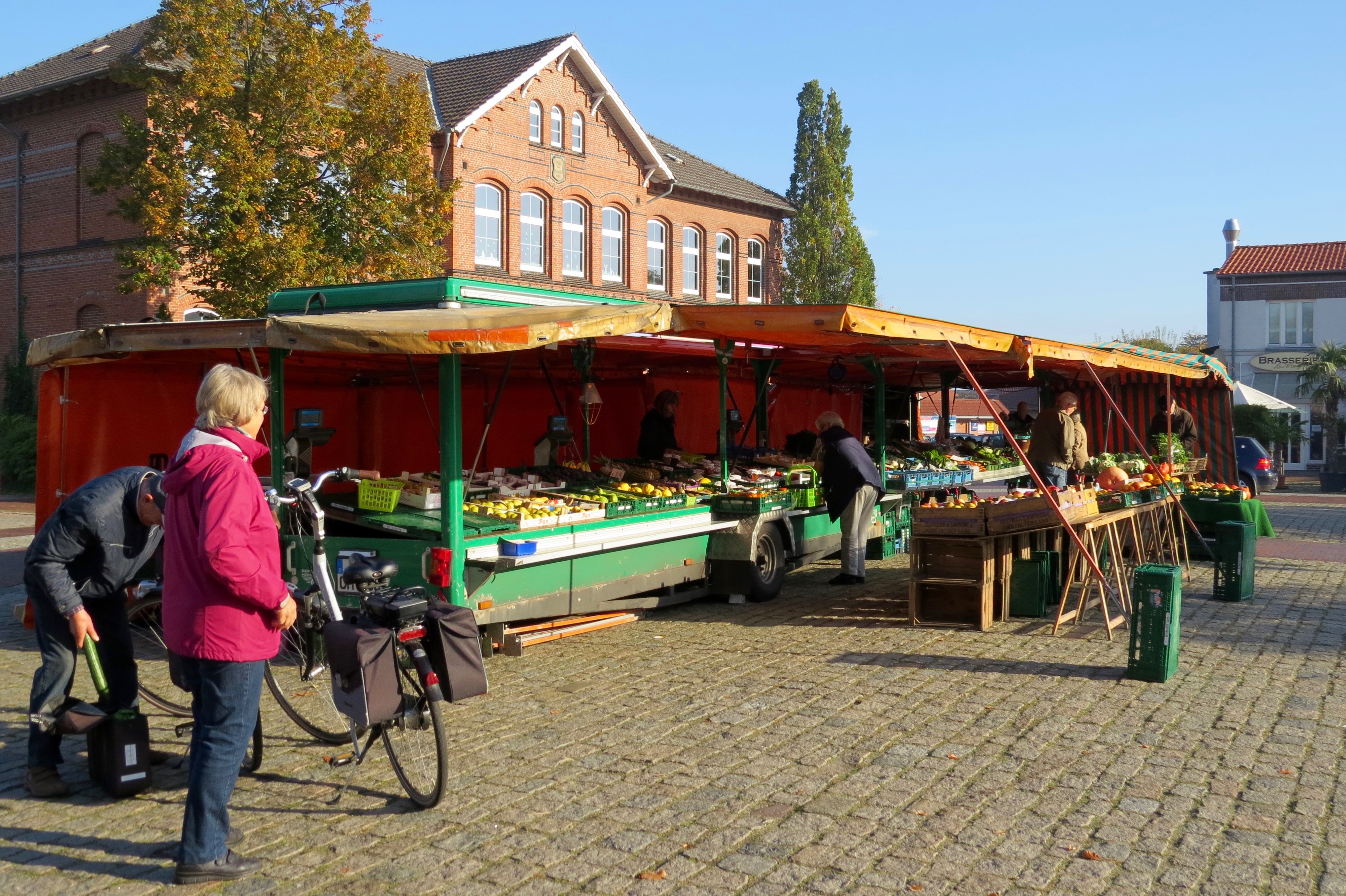 Marktstand mit Obst und Gemüse auf einem öffentlichen Platz mit Marktbesuchern. Im Hintergrund ein großes rot geklinkertes Gebäude.