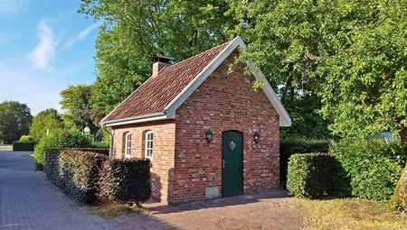 Ein Backhaus aus rotem Ziegelstein mit Satteldach, umgeben von grünen Bäumen und roten Sträuchern
