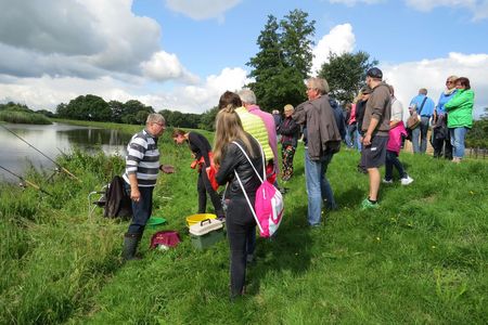 Eine Menschengruppe steht an einem Fluss mit belaubtem Hintergrund. Ein Angler gibt de Gästen eine Erklärung dazu.