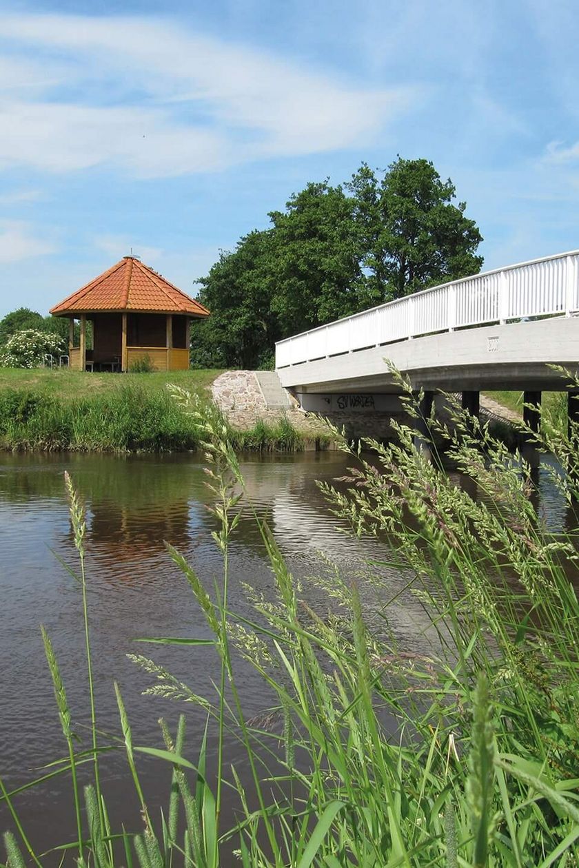 Eine weiße Brücke überspannt einen Fluss. Im Hintergrund steht eine Schutzhütte aus Holz mit rotem Dach