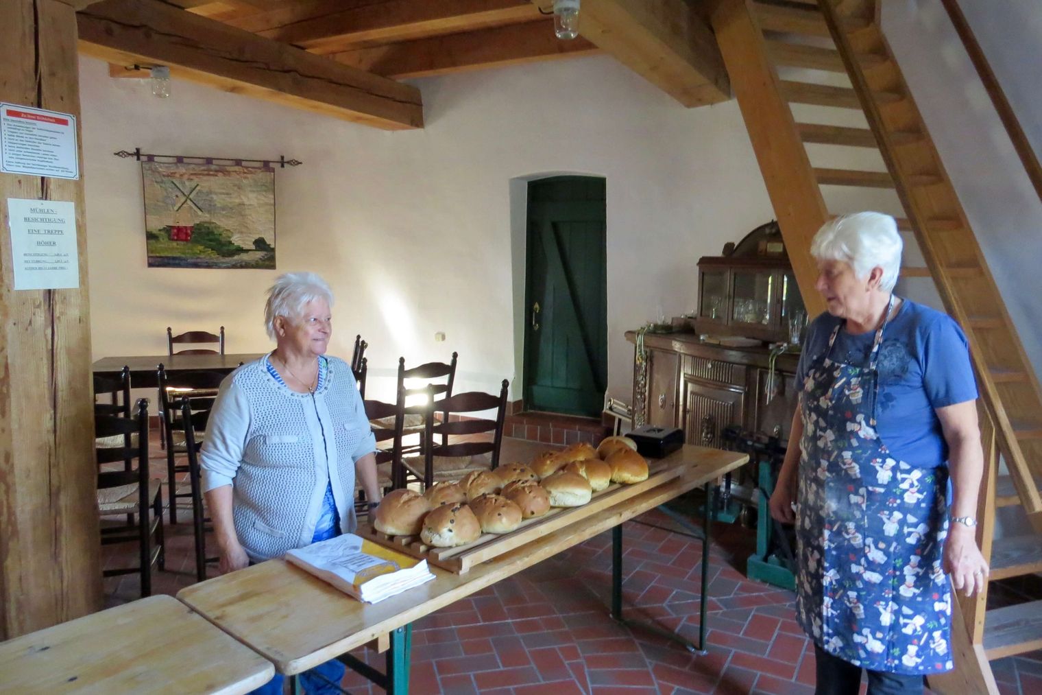 2 Frauen verkaufen frisch gebackenen Stuten auf Holztischen. Im Hintergrund ein, mit alten Möbeln eingerichteter Raum.