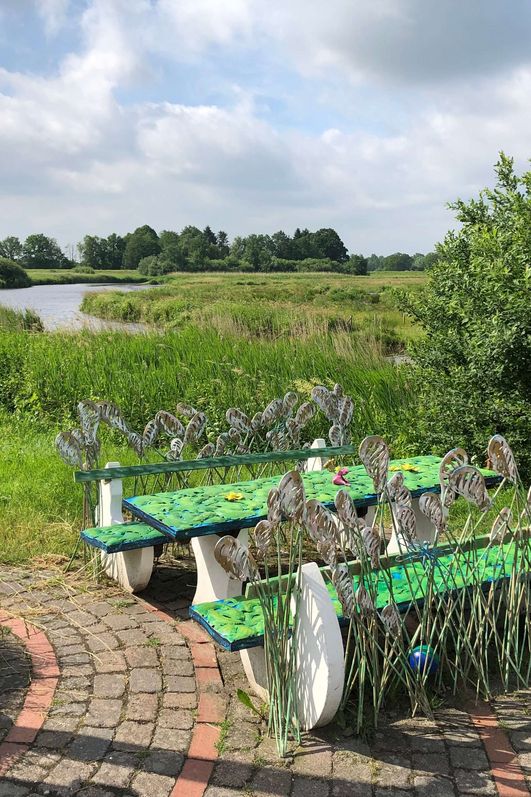 Eine verschiedenfarbige Eisenbank steht auf einem grünen Deich mit Blick auf einen Fluss