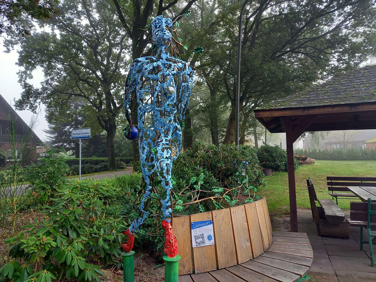 Ein Schild mit einem QR Code ist an einer Holzbank befestigt. Daneben eine bunte Skulptur aus Eisen.