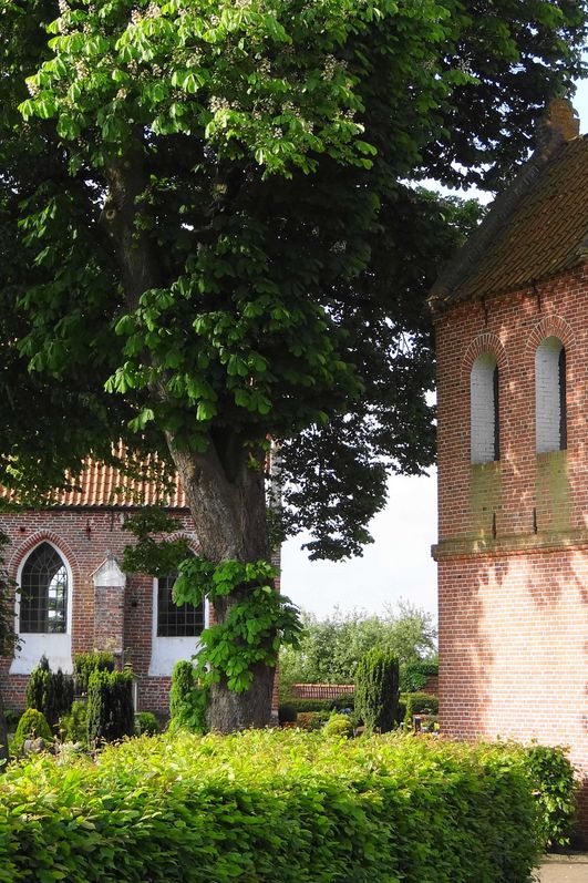 Hinter belaubten Bäumen und einer Hecke ist eine kleine Kapelle auf einer Warft zu sehen. Im Vordergrund befindet sich ein freistehender Glockentrum mit Durchlass für Besucher.