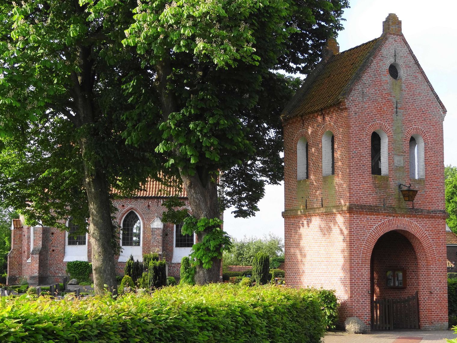 Hinter belaubten Bäumen und einer Hecke ist eine kleine Kapelle auf einer Warft zu sehen. Im Vordergrund befindet sich ein freistehender Glockentrum mit Durchlass für Besucher.