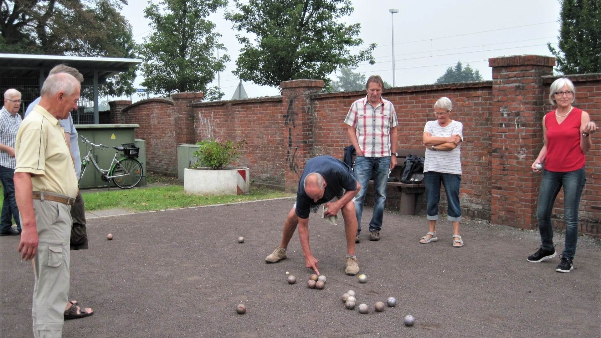 Eine Gruppe aus Frauen und Männern spielt Boule auf einem sandigen Platz. Ein Mann steht in gebeugter Haltung und und zeigt auf zwei silberne Kungeln.