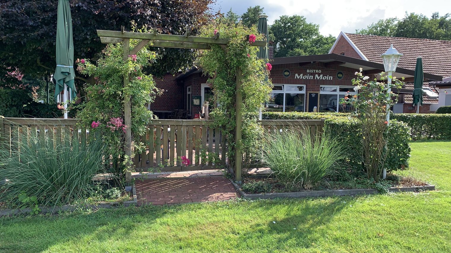 Hölzernes Gartentor mit Rankpflanzen (Rosen) und viel Grün, im Hintergrund ein Backsteingebäude mit der Aufschrift „Bistro Moin Moin“