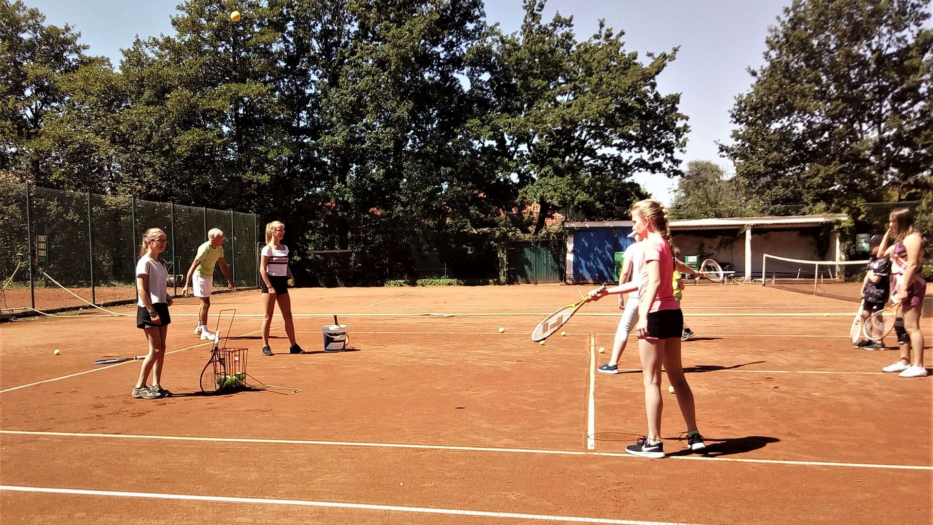 Ein Tennisplatz mit rötlichen Sand im Sonnenschein. Mehrere Kinder versuchen sich im Tennisspiel.