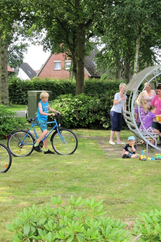 Auf einer begrünten Rasenfläche mit belaubtem Hintergrund befindet sich eine Personengruppe beim picknicken auf einem Rastplatz. Im Vordergrund stehen zwei Jungen mit ihren Fahrrädern. 
