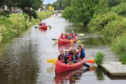 Mehrere Paddelboote auf einem Kanal mit Paddlern in Schwimmwesten legen ab vom Ufersteg. Der Kanal ist eingefasst von grüner Uferböschung.