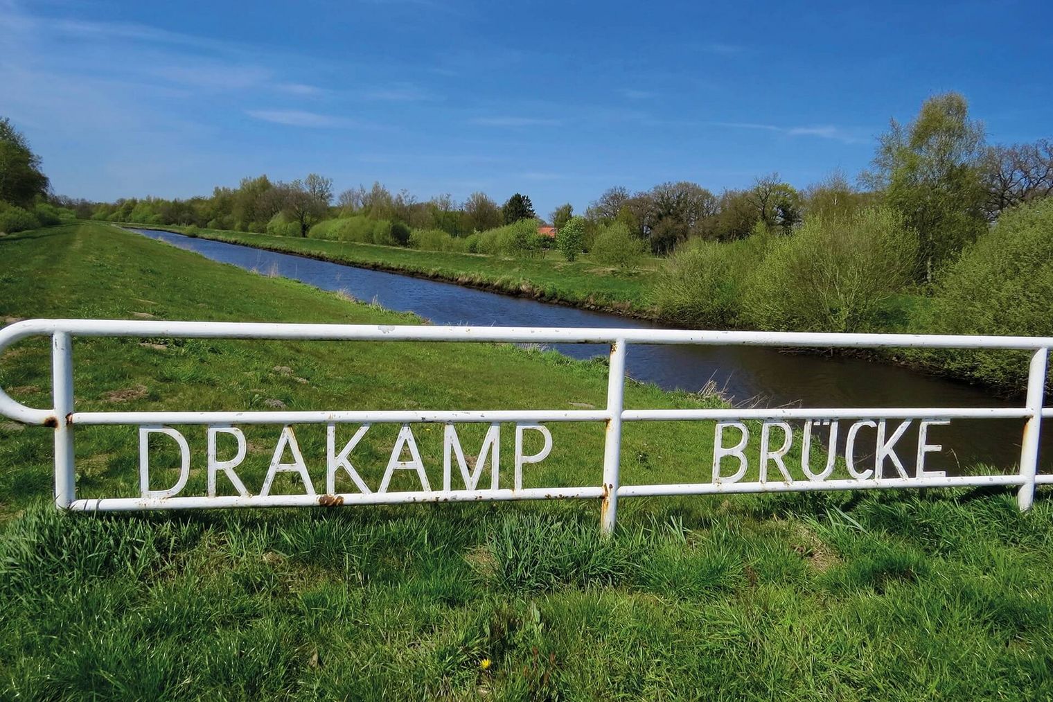 Ein weißes Brückengeländer mit Einschrift über einem Fluss. Im Hintergrund belaubte Bäume und grüne Wiesen