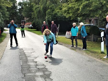Eine Frau spielt eine Boßelkugel auf einer Straße.Im Hintergrund beobachten und fotografieren die Mitspieler