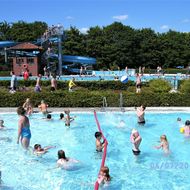 In einem Schwimmbecken mit niedriger Wassertiefe sind mehrere Personen in Badekleidung zu sehen. Im Hintergrund ein großes Schwimmbecken mit einem Turm und einer Wasserrutsche.