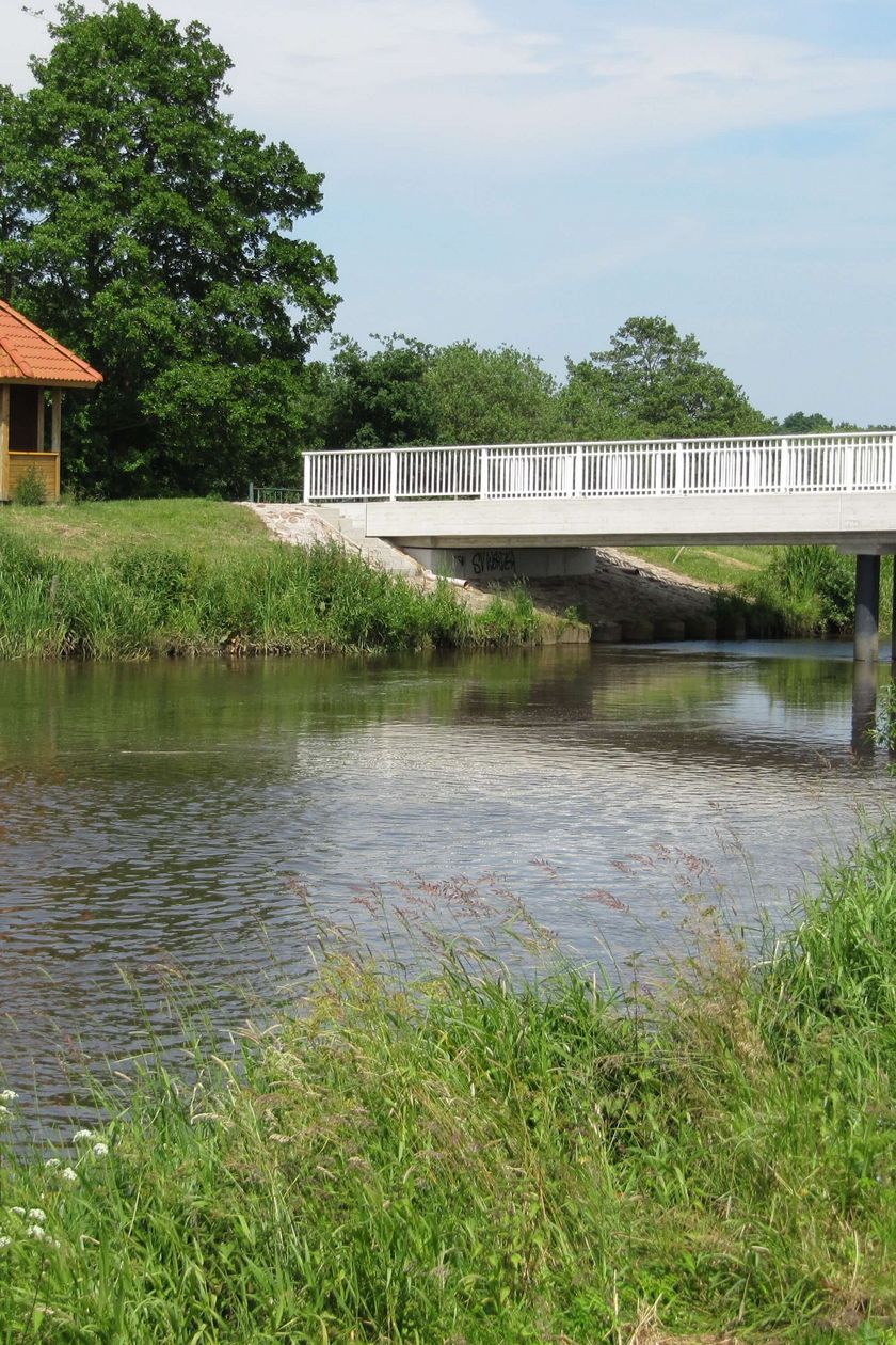 Schutzhütte am Uferrand eines Flusses. In unmittelbarer Nähe überspannt eine weiße Brücke den Fluss. Im Hintergrund ist eine grüne Landschaft mit Bäumen zu sehen.