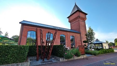 Backsteinindustriegebäude mit Turm und großen Fenstern vor klarem Himmel; moderne Kunstinstallation vorne.