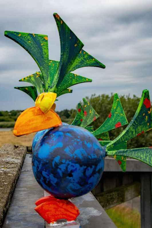 Bunte Vögel aus Stahl sitzen auf einemHolzverschlag. Im Hintergrund ist eine Deichlandschaft