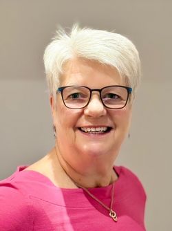 Porträt einer lächelnden Frau mit kurzen grauen Haaren, Brille und pinkfarbenem Oberteil.