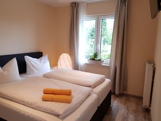 Gemütliches Doppelzimmer mit zwei Einzelmatratzen, hellen Vorhängen und einem Fenster mit Blick ins Grüne. Auf dem Bett liegen zwei gefaltete, orangefarbene Handtücher, daneben steht eine moderne Lampe und eine Blumendekoration auf der Fensterbank.