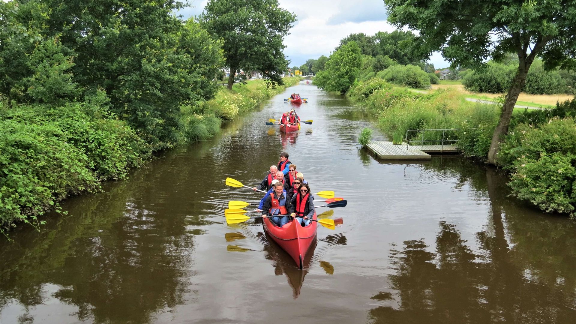 Mehrere Paddelboote mit Paddlern in Schwimmwesten fahren auf einem Kanal mit grüner Böschung am Ufer.