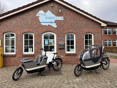 Zwei moderne Lastenfahrräder der Marke Gazelle parken vor der Apen Touristik, ein Gebäude mit weißer Tür und weißen Fenstern.
