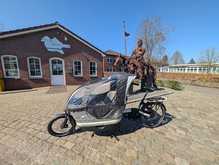 Ein Lastenrad, platziert auf einem gepflasterten Hof vor der Apen Touristik, daneben eine Skulptur einer historischen Figur