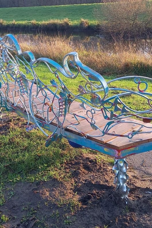 Eine verschiedenfarbige Eisenbank steht auf einem grünen Deich mit Blick auf einen Fluss