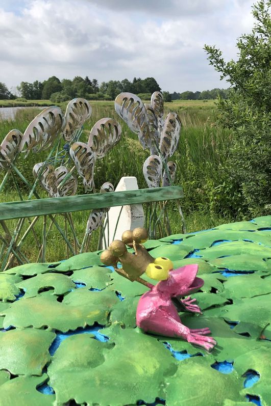 Ein rosa Froschkönig aus Metall sitzt auf einem Blättertisch aus Metall.