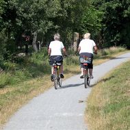 Das Foto zeigt ein 2 ältere Damen auf dem Fahrrad von hinten. Sie fahren auf einem geschotterten Fahrradweg zwischen grünen Wiesen. Im Hintergrund belaubte Bäume.